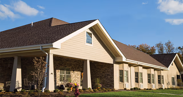 Front exterior of a single-story senior living building with gabled roofs, stone and siding facade, columns and a landscaped lawn.