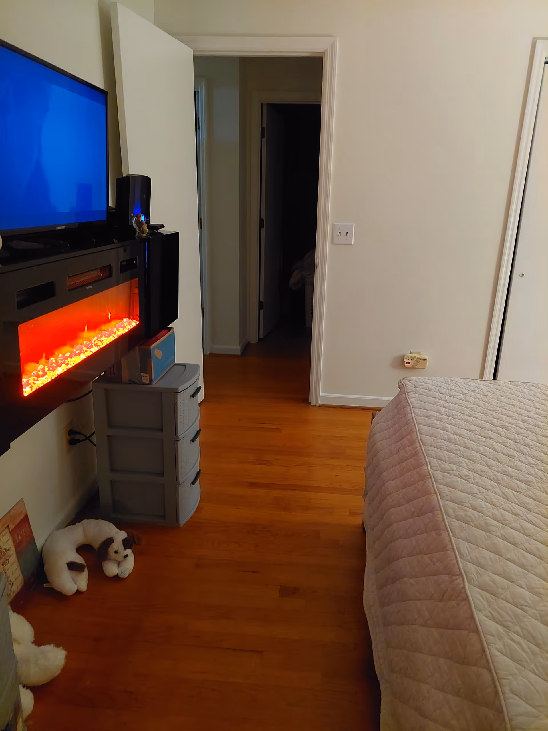 Cozy bedroom with bed, electric fireplace, TV, and stuffed toy on wooden floor.