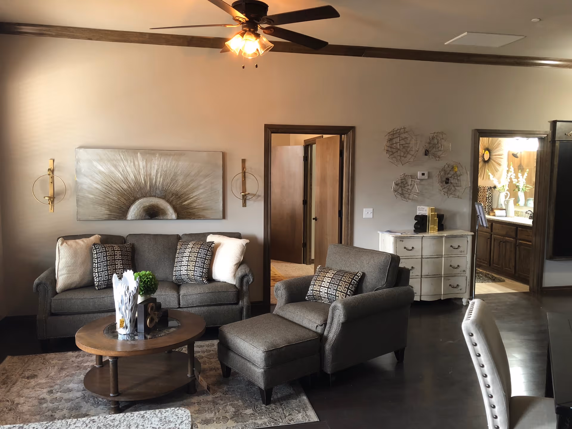 A cozy living room with a gray sofa and matching armchair with ottoman, decorated with patterned and plain cushions. A round wooden coffee table with decorative items sits on a patterned rug. The walls are beige with wooden trim, featuring a large abstract artwork and two wall sconces. A ceiling fan with lights is mounted on the ceiling. In the background, there is a doorway leading to another room and a glimpse of a bathroom with a large mirror and floral decor.