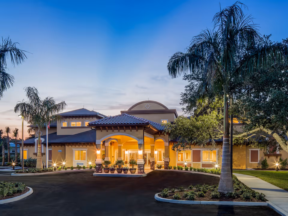 Exterior view of Aravilla Sarasota senior living facility at dusk, showing a well-lit entrance with arches, palm trees, landscaped garden beds, and a clear sky.