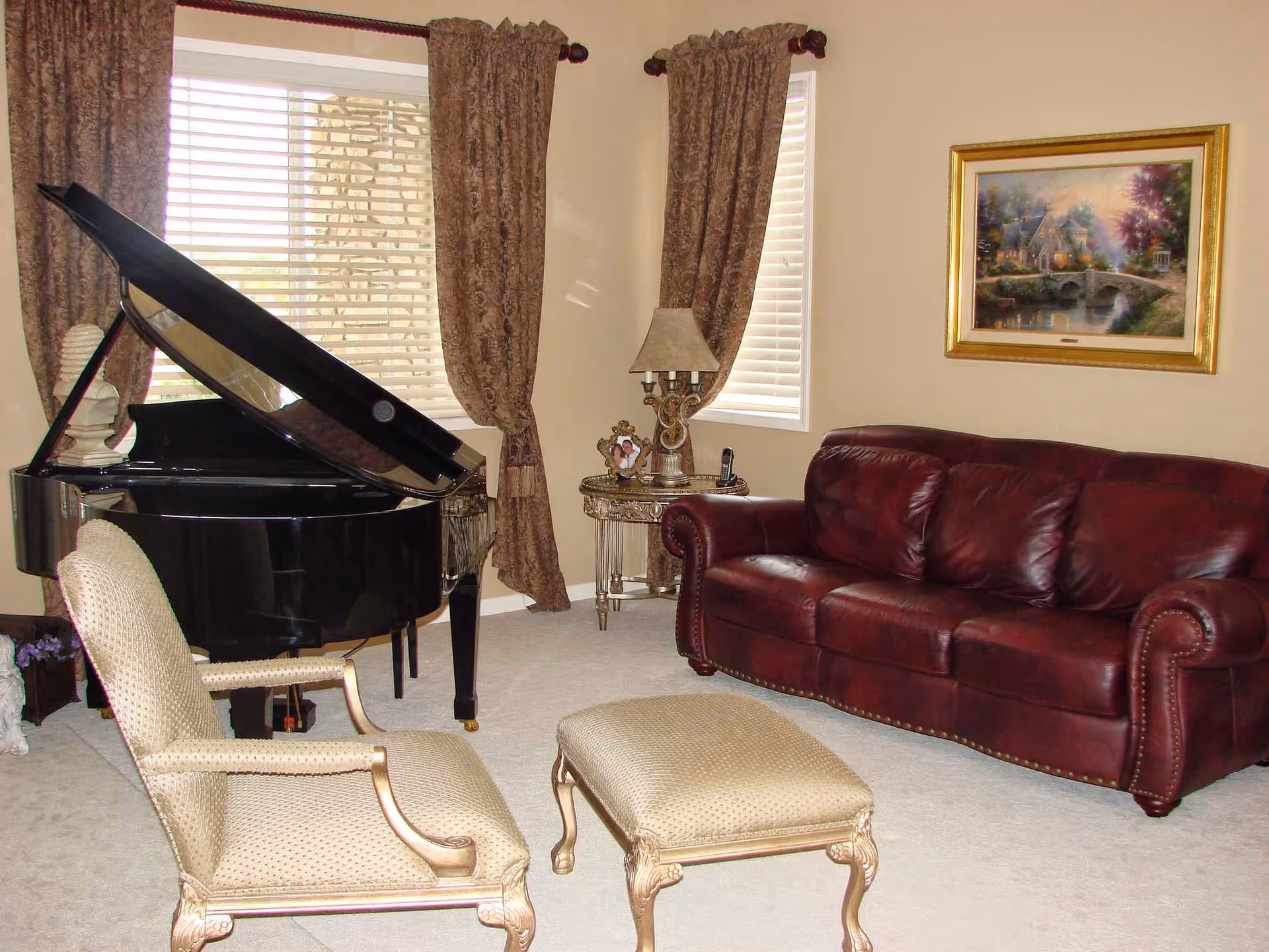 A cozy living room featuring a grand piano, burgundy sofa, upholstered chairs, and draped curtains.