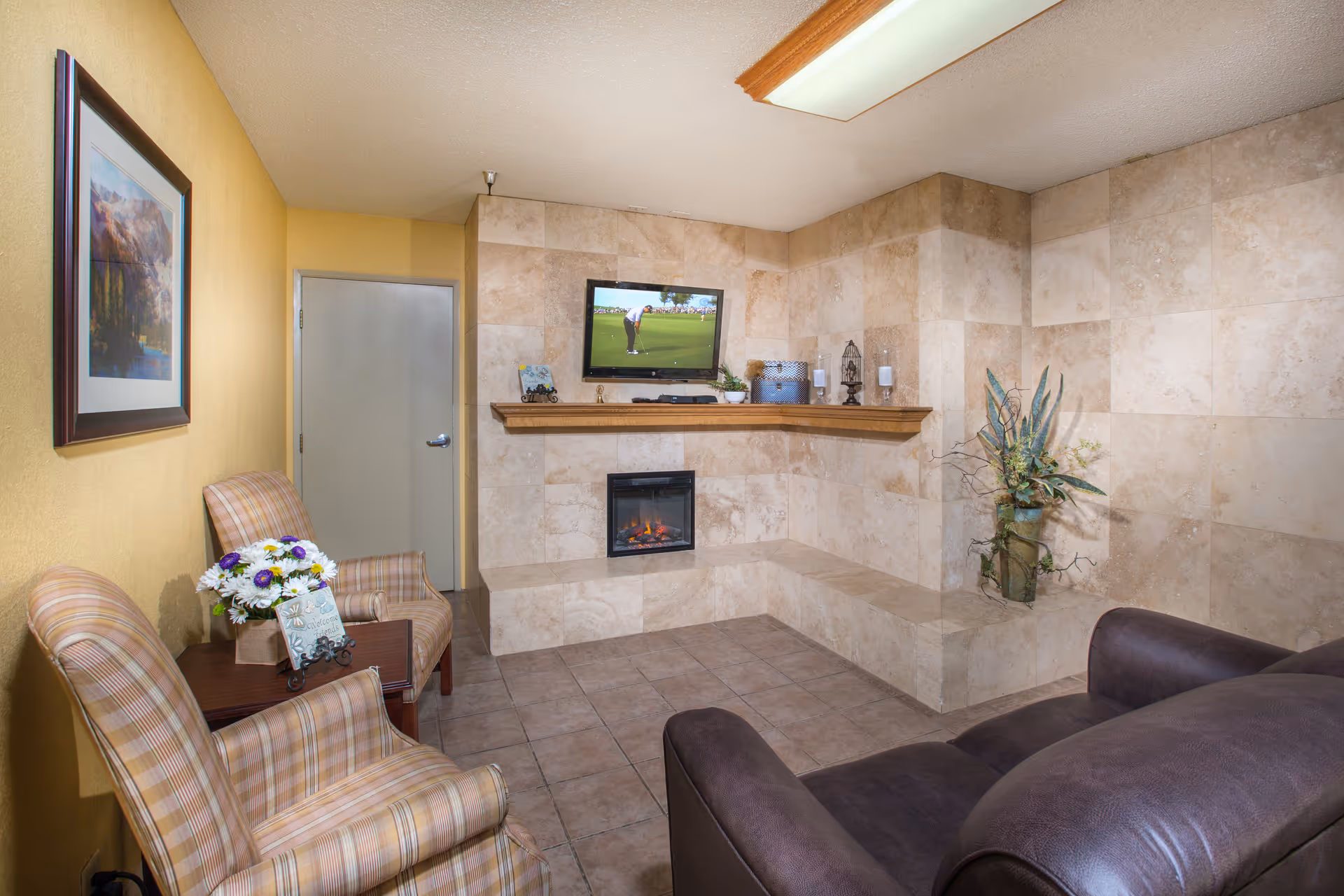 A cozy living room area with a tiled fireplace and a mounted flat-screen TV showing a golf game. The room has beige tiled walls and floor, a brown leather sofa, two plaid armchairs, a small wooden side table with a flower arrangement and a decorative sign, and a framed landscape painting on a yellow wall.