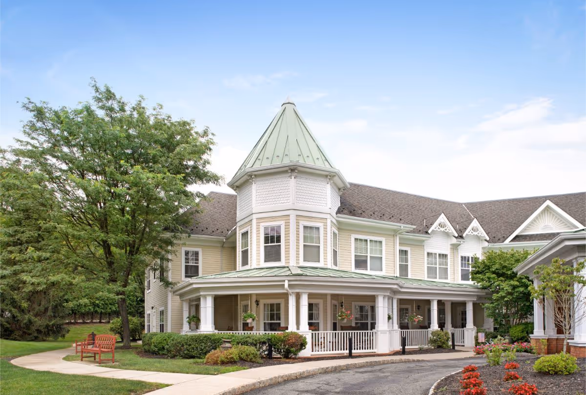 Front exterior of a large Victorian-style senior living building with a wraparound porch, turret, and landscaped grounds.