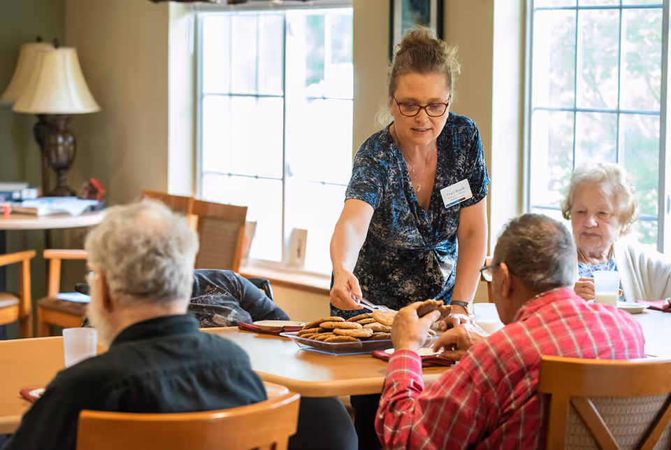 A staff member serves cookies to elderly residents seated around a dining table by large windows.