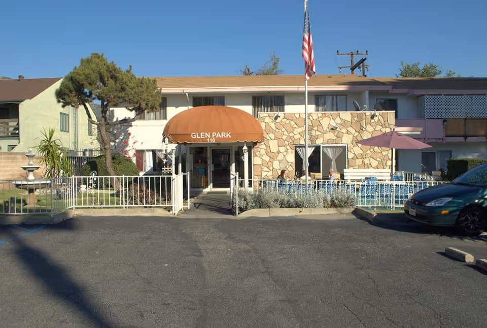 Front entrance of a two-story senior living building with an orange awning labeled "GLEN PARK", flagpole, fountain, and outdoor seating.