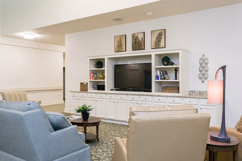 A cozy living room area with a large white entertainment center holding a flat-screen TV, decorative plants, books, and baskets. In front of the entertainment center are comfortable chairs, a small round wooden table with a potted plant, and a floor lamp with a pinkish shade. The walls are white with framed botanical prints and a decorative wall piece.