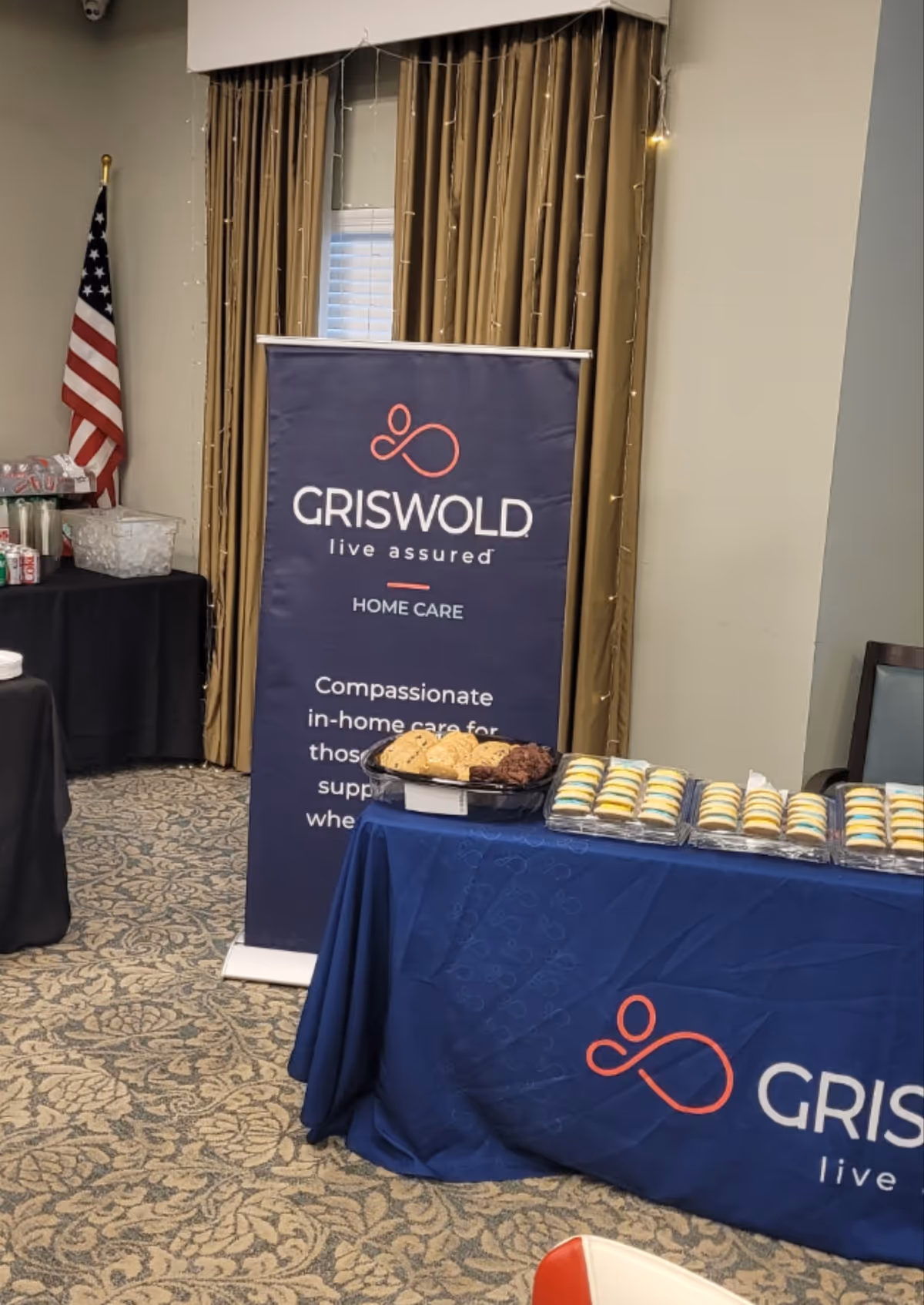 Indoor event setup featuring a Griswold Home Care banner, cookies, and a flag.