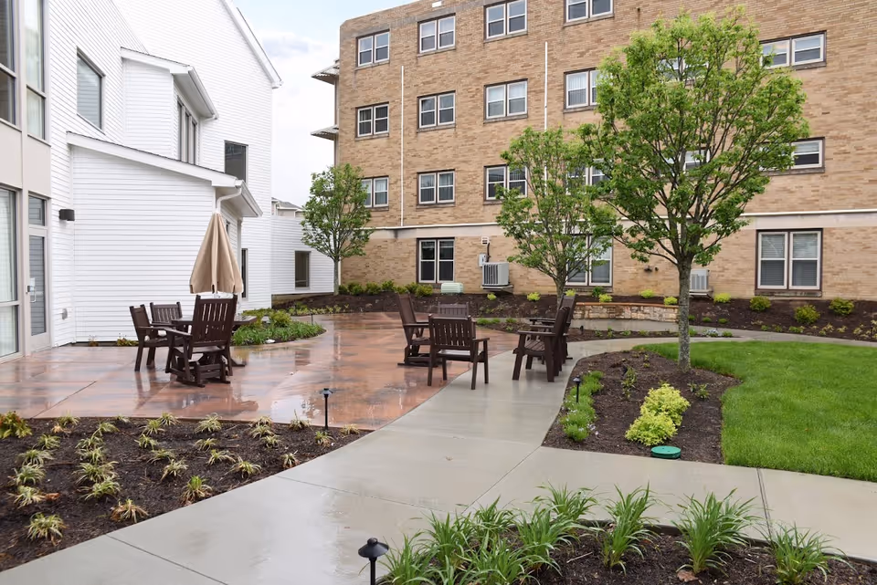 Outdoor courtyard with wooden tables and chairs, trees, and a brick residential building in the background.