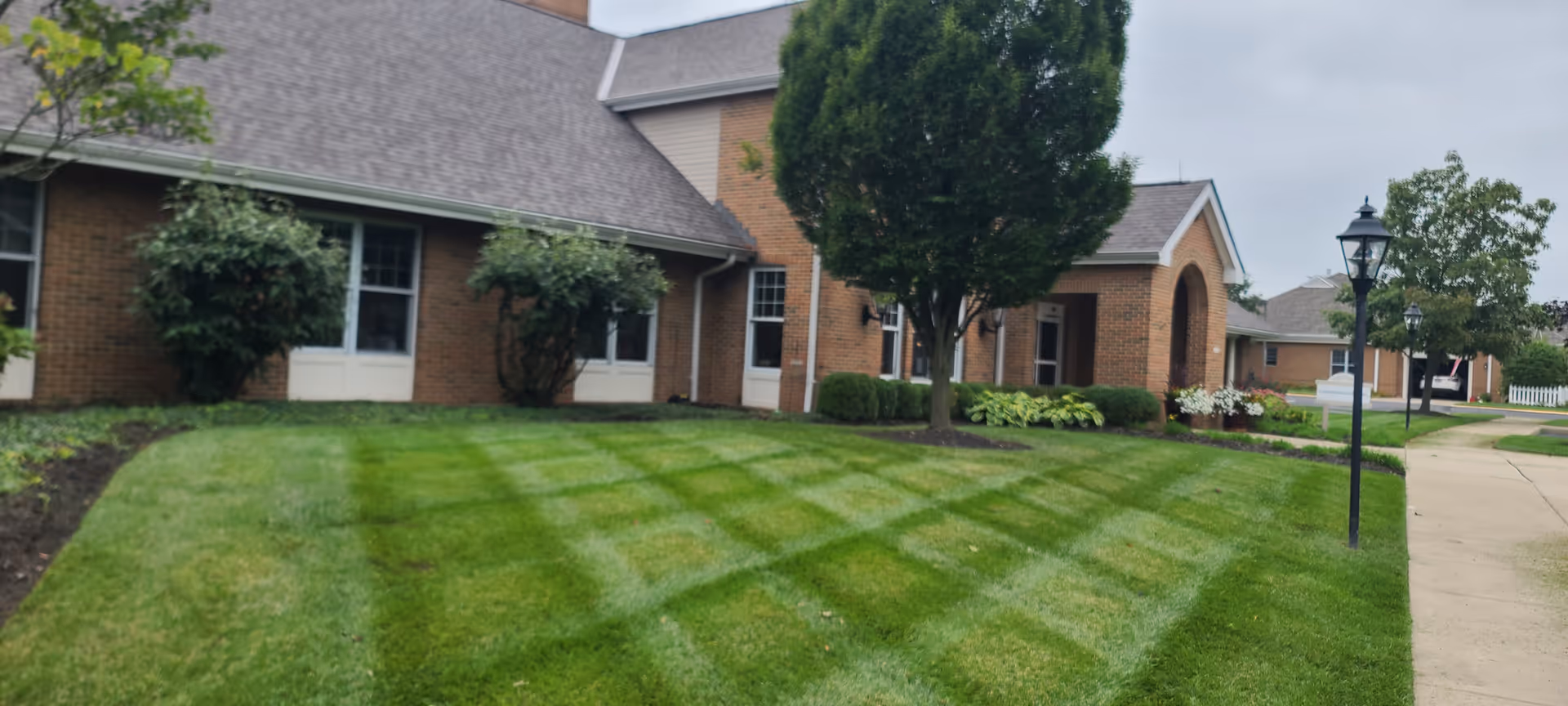 Brick senior living facility with manicured lawn, trees, and walkway along its broad front facade.