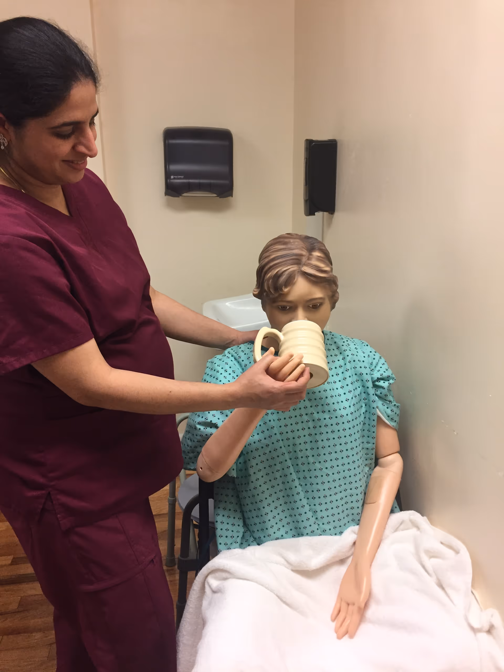 Caregiver in maroon scrubs assists a mannequin with a mug in a clinical training room.