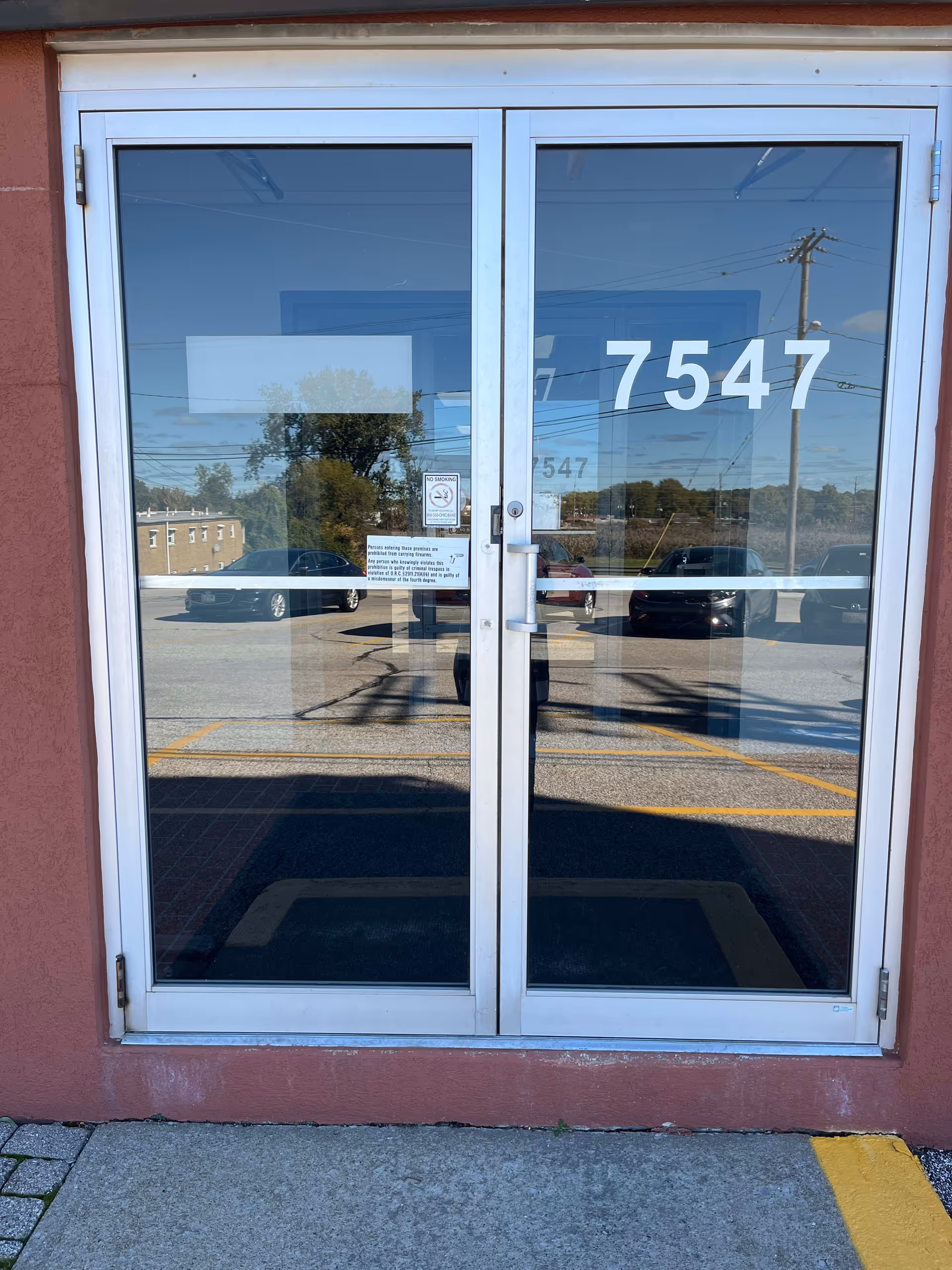 Double glass doors with white frames on a red exterior wall, reflecting parking lot and street.