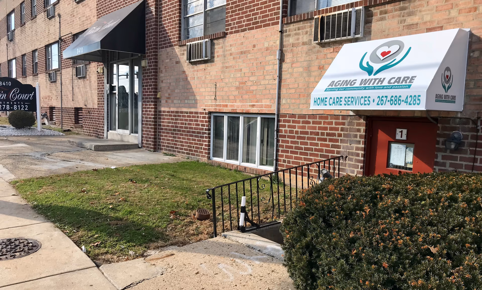 Brick exterior with Aging With Care awning, entrance stairs, and surrounding shrubs.