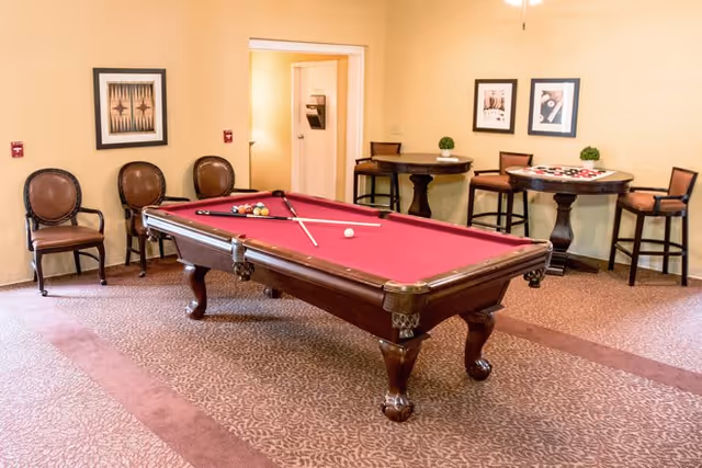A recreational room with a red felt pool table in the center, pool cues and balls arranged on it. The room has beige walls, patterned carpet, and several chairs along the walls. Two small round tables with chairs and decorative plants are placed against the far wall, which also features framed artwork.