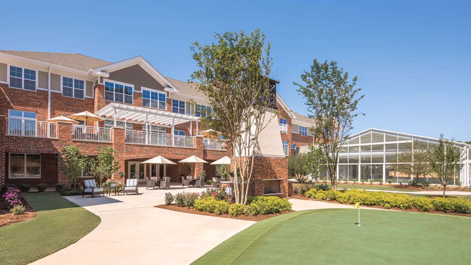 Brick three-story senior living building with a shaded outdoor patio, seating areas, landscaped grounds and a small putting green.