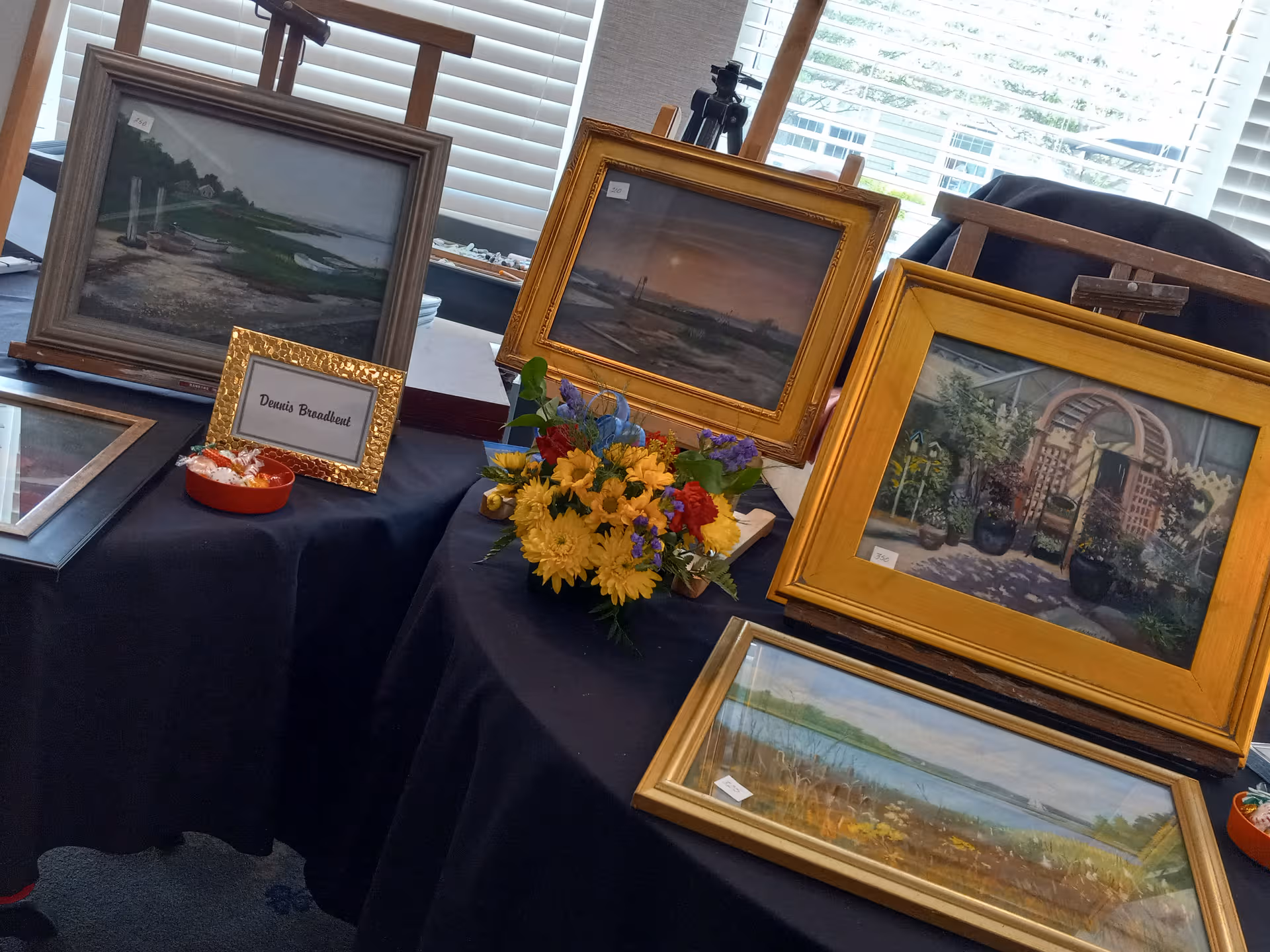 Framed landscape paintings displayed on tables with a bouquet indoors.