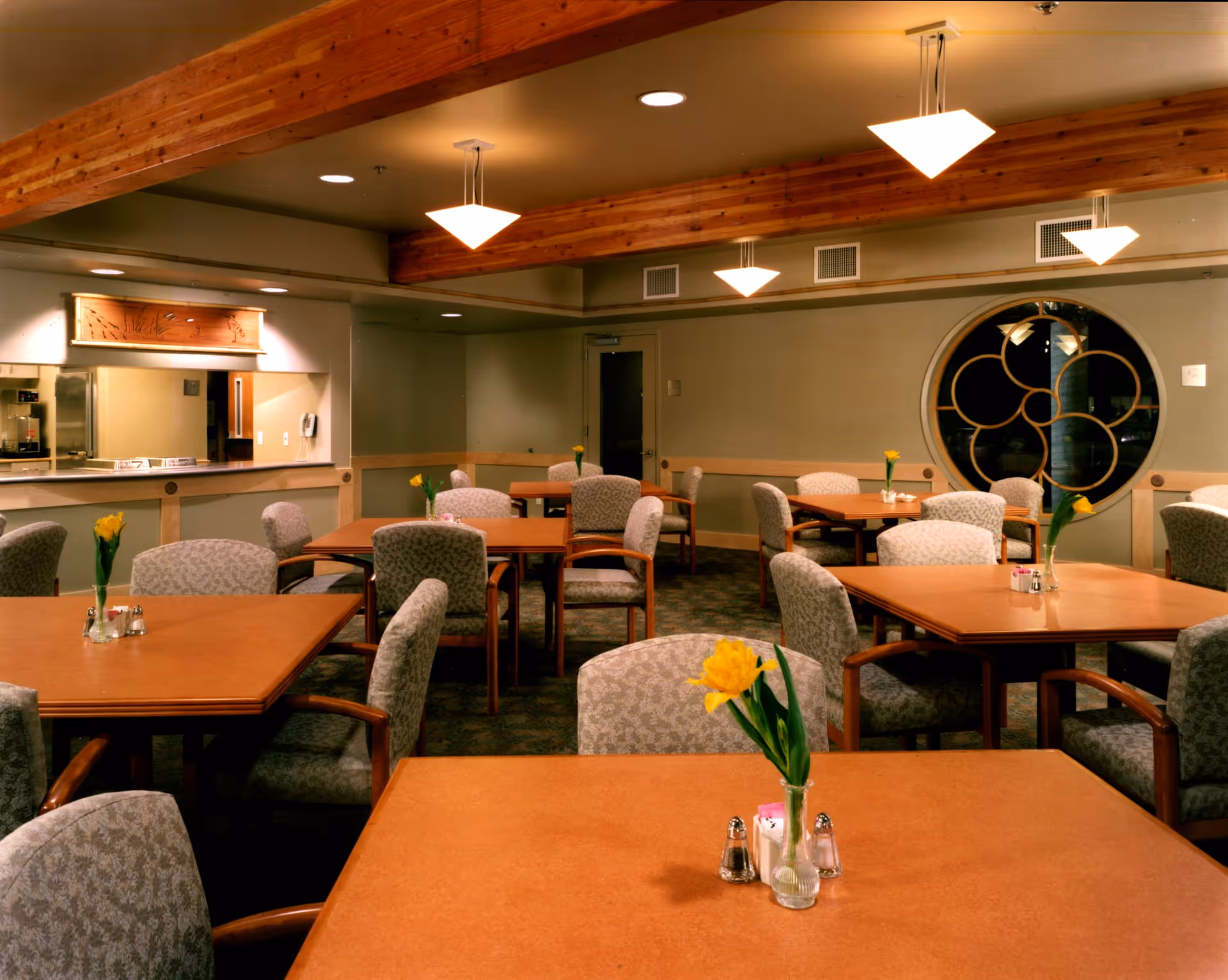 Dining room with multiple wood tables and upholstered chairs, each table topped with a vase holding a yellow flower.