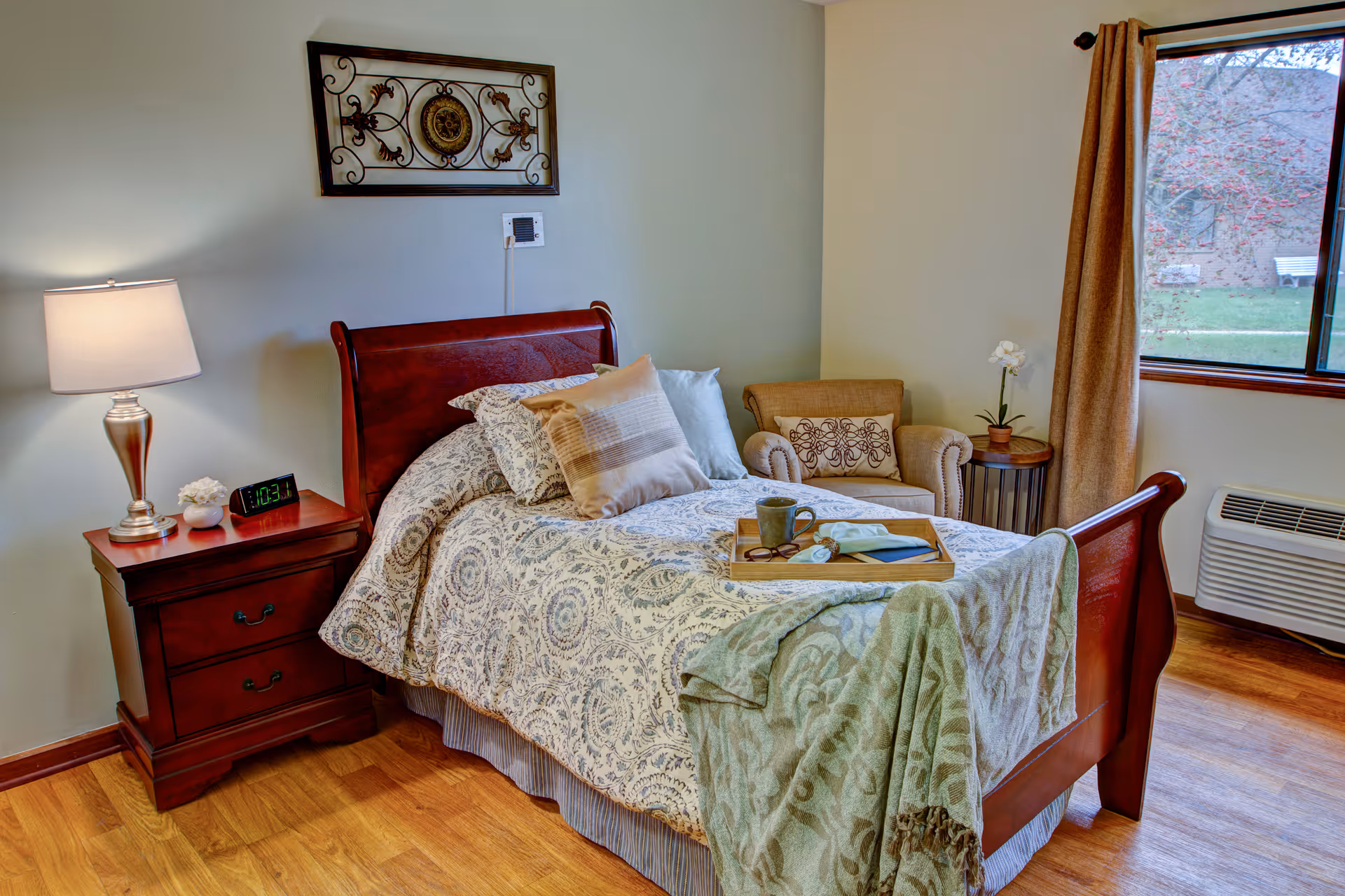 A cozy bedroom with a wooden four-poster bed, nightstand, lamp, and seating by a window.