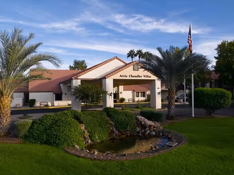 Front view of Atria Chandler Villas exterior with manicured lawn, palm trees, and a pond.