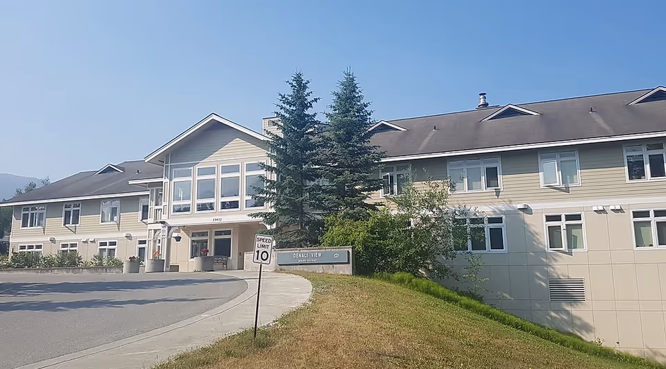 Front exterior of a two-story senior living building with large windows, evergreen trees, a curved driveway, and a speed limit sign.