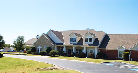 Exterior view of a single-story senior living facility building with a combination of brick and siding, a covered entrance, and a paved driveway surrounded by green lawns and some trees under a clear blue sky.