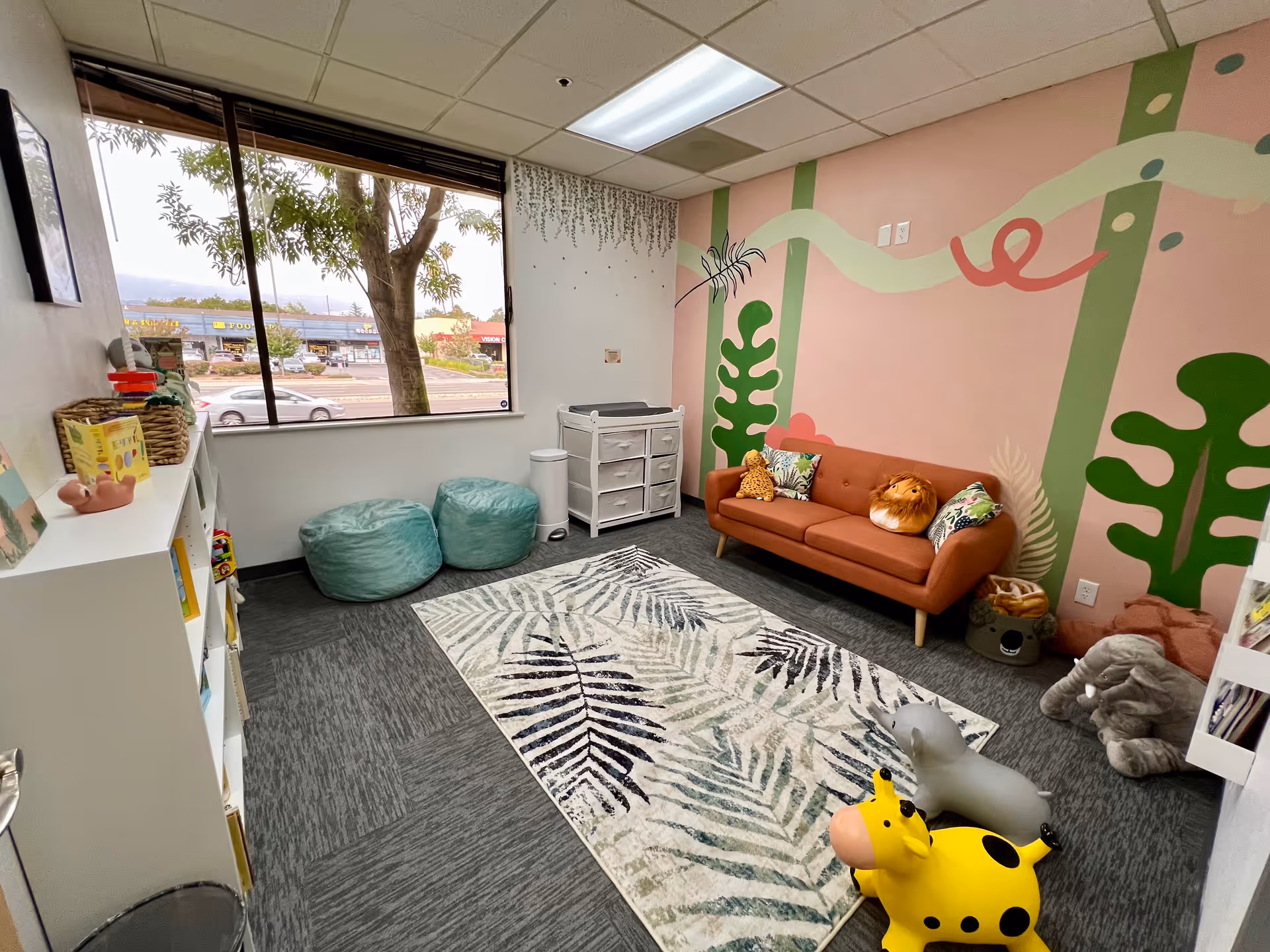 Colorful children's therapy room with pink mural wall, sofa, rug, shelves, and stuffed toys.