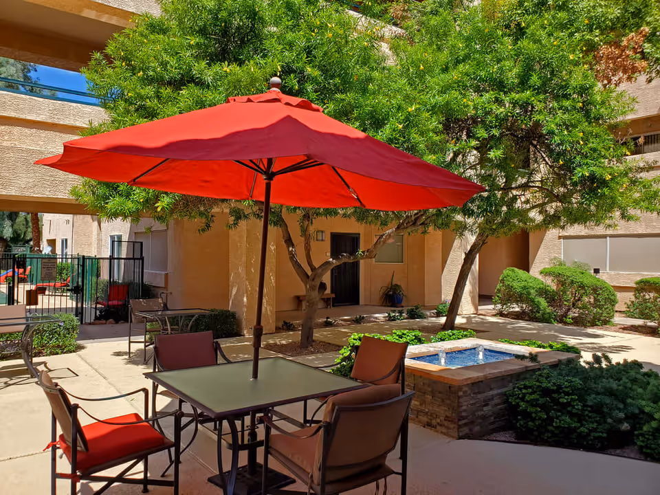 Outdoor patio area with a square table and four chairs, each with red cushions, shaded by a large red umbrella. Surrounding the patio are green trees and bushes, with beige building walls in the background and a small water feature nearby.