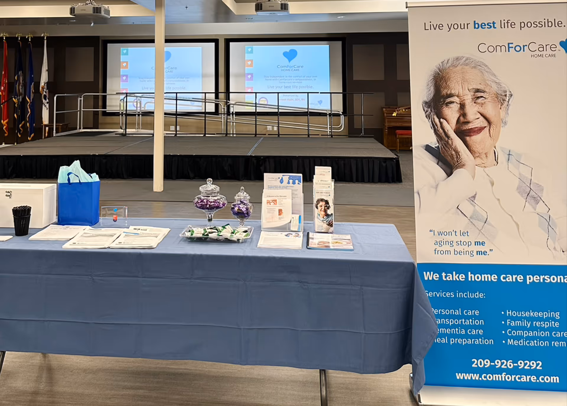 Indoor event setup with a blue-clothed table, banners, and a stage in a senior care facility.
