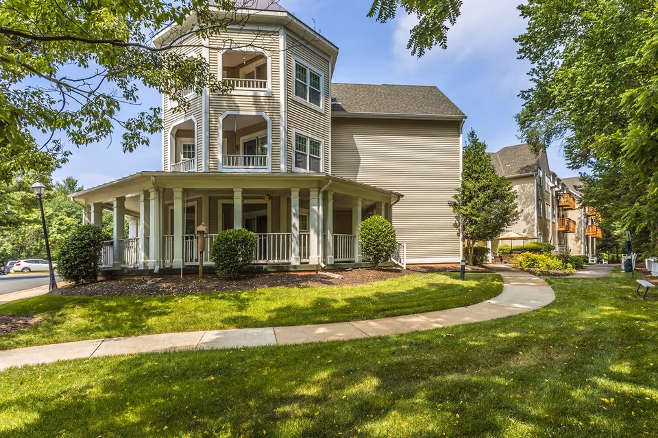 Exterior view of a multi-story senior living building with a wraparound porch, balconies, and a curved sidewalk through a landscaped lawn.