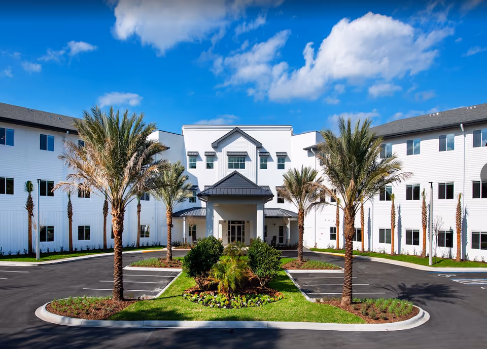 Front exterior view of a large, white, three-story assisted living and memory care facility with a covered entrance, palm trees, landscaped greenery, and a parking area under a blue sky with scattered clouds.