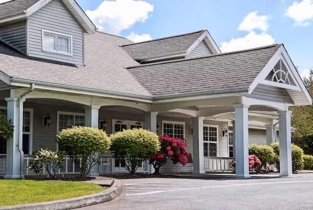 Front entrance of a light-gray senior living building with a covered porte-cochère, white columns, landscaped shrubs and pink flowering bushes.