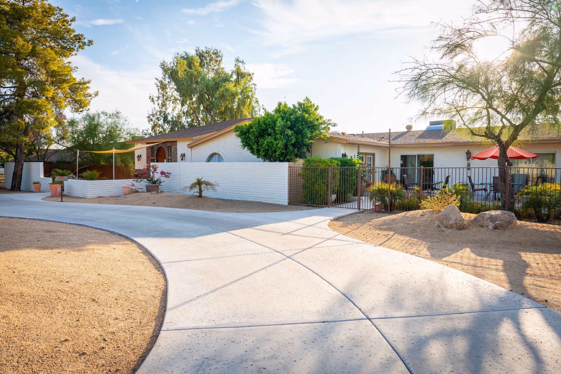 Front exterior of a single-story care home with a curved driveway, white brick wall, patio area and desert landscaping.