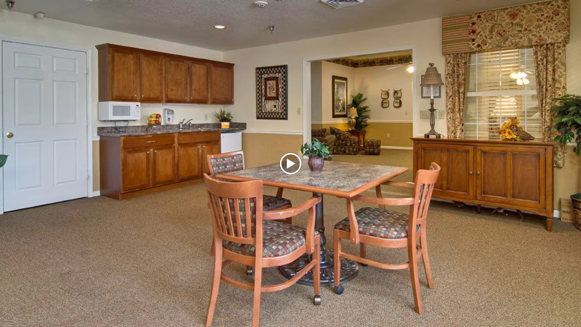 A cozy senior living facility common area featuring a small dining table with four wooden chairs with patterned cushions. To the left, there is a kitchenette with wooden cabinets, a microwave, a sink, and a dishwasher. In the background, there is a living room area with a sofa, plants, and framed artwork on the walls. A wooden sideboard with decorative items and floral curtains adorn the window on the right side.
