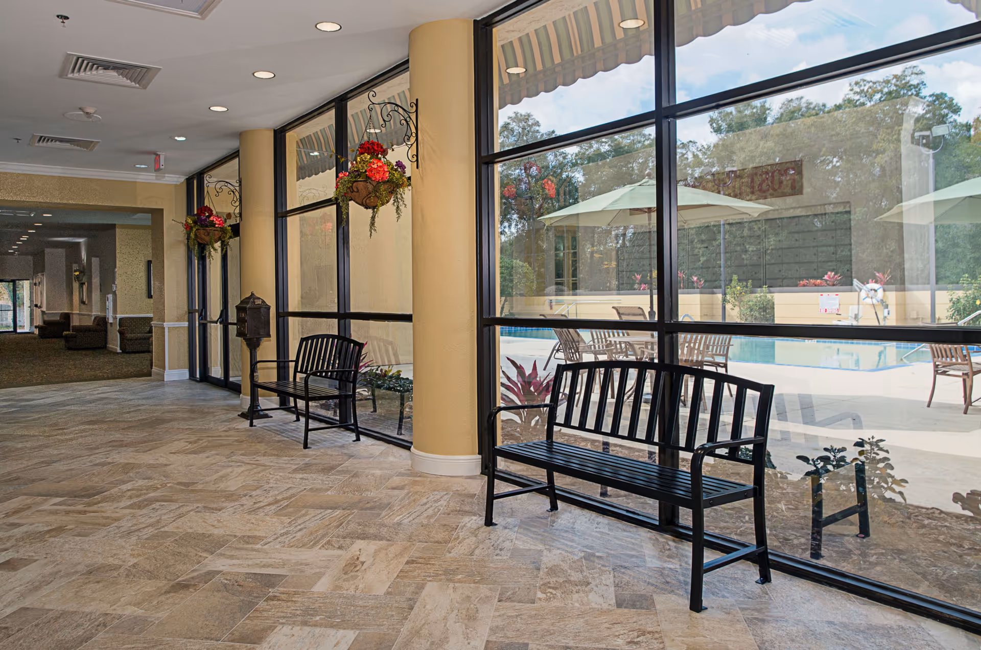 Indoor lounge with large windows, black benches, hanging flowers, and pool view outside.