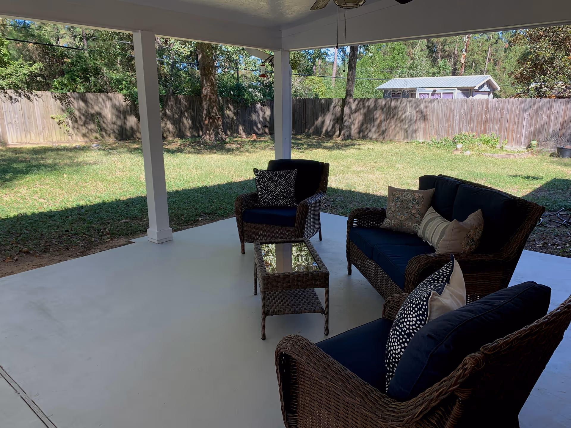 Covered patio with wicker seating and glass coffee table overlooking a grassy backyard.
