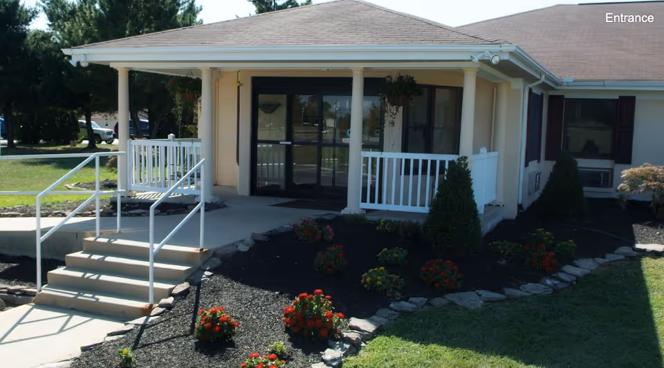 Front entrance of a single-story building with a covered porch, steps, railing, and landscaped flower beds.