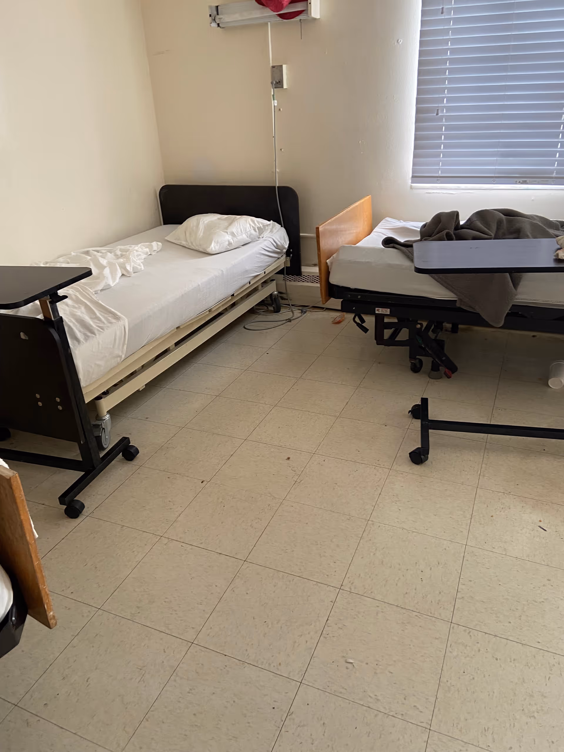 Two hospital beds in a small patient room with a window and minimal furnishings.