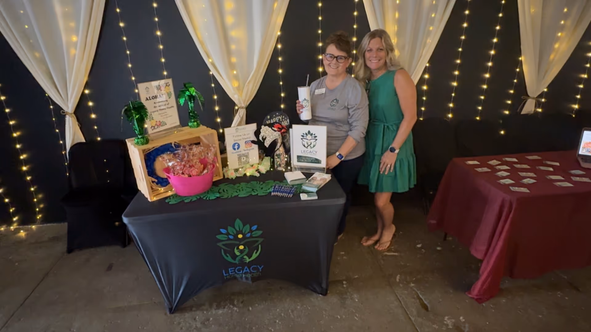 Two women stand beside a promotional booth with a Legacy Home Health logo and string lights.