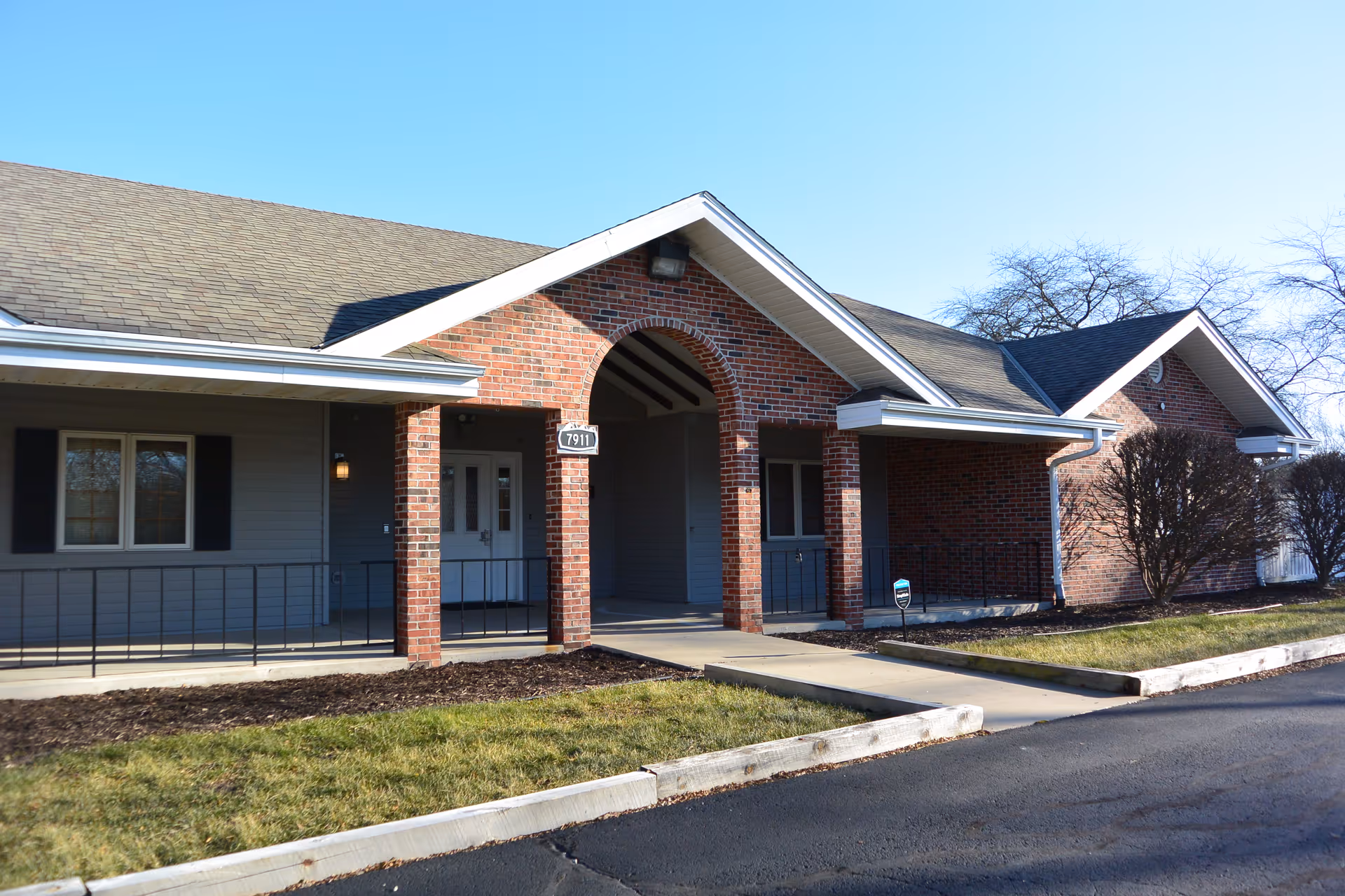 Exterior view of a single-story brick and siding building with a covered entrance supported by brick columns. There is a sidewalk leading to the entrance, some grass and landscaping in front, and a clear blue sky above.