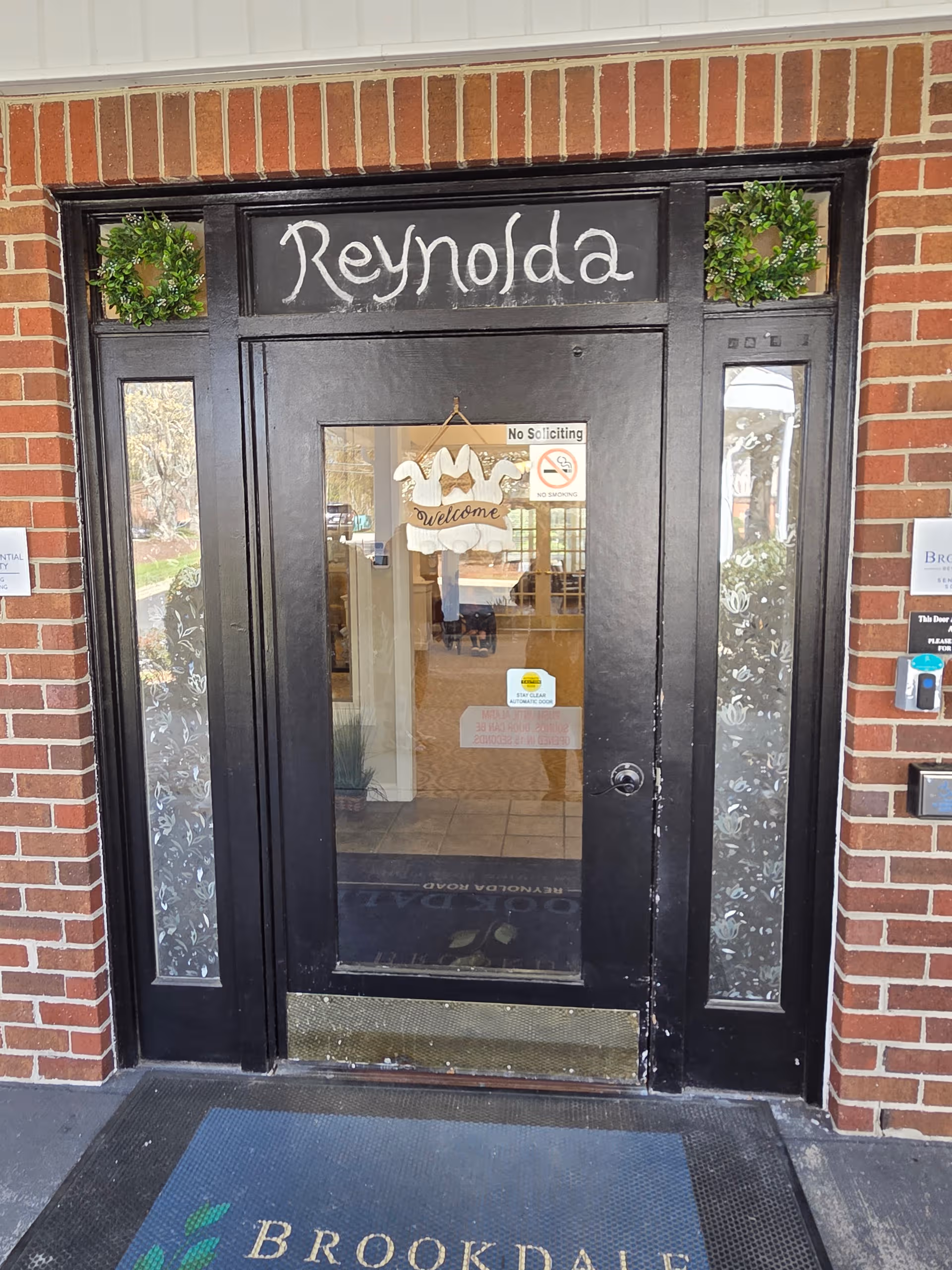 Black front door with wreaths and Brookdale Reynolda Road signage at a brick entrance.