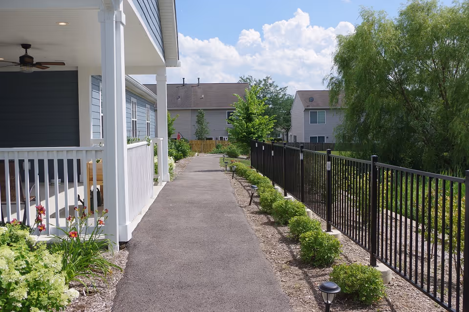 A paved walkway runs beside a covered porch with hedges, a black fence, and trees.
