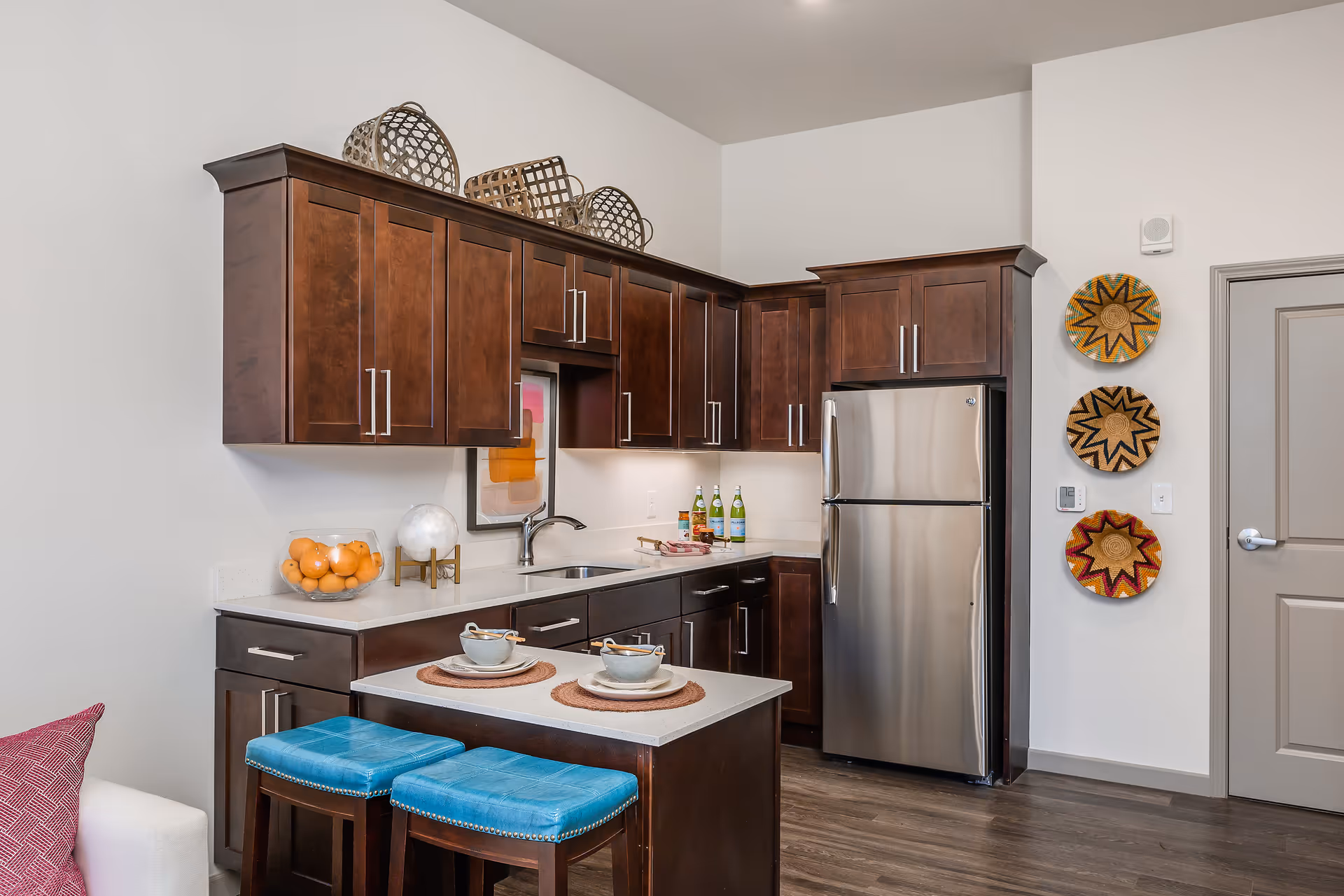 Modern kitchen area with dark wooden cabinets, a stainless steel refrigerator, a small island with two blue cushioned stools, and decorative baskets on the wall and above the cabinets. The countertop has a bowl of oranges, a small globe, and two place settings with cups and plates.