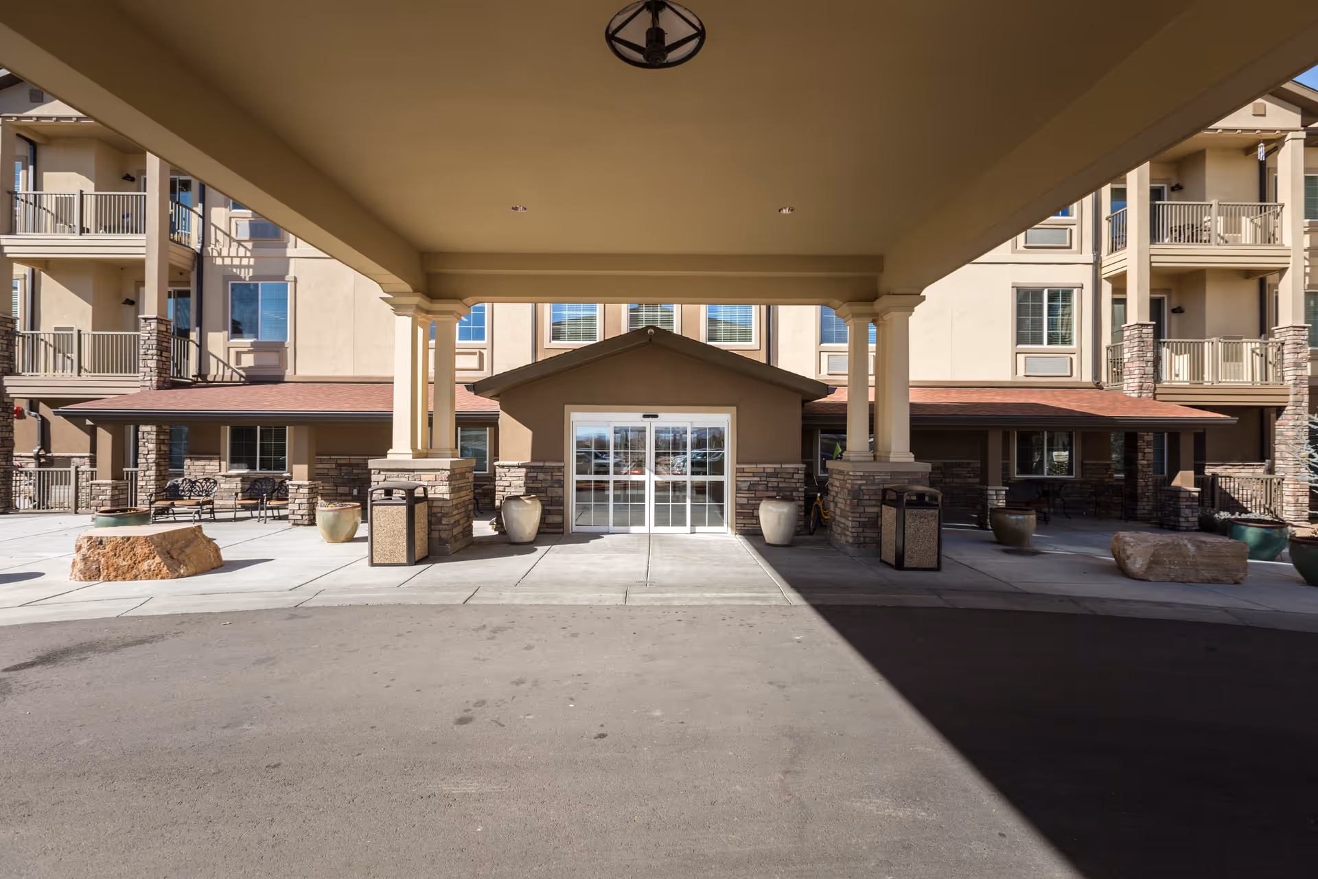 Entrance area of a senior living facility with a covered drop-off zone supported by columns. The building has multiple floors with balconies and stone accents. There are large planters and benches near the entrance, and automatic sliding glass doors lead inside.