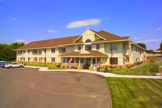 Exterior view of a two-story assisted living facility building with a beige and brown color scheme, a peaked roof, and a covered entrance. There are a few cars parked in the parking lot, green grass, and a clear blue sky.