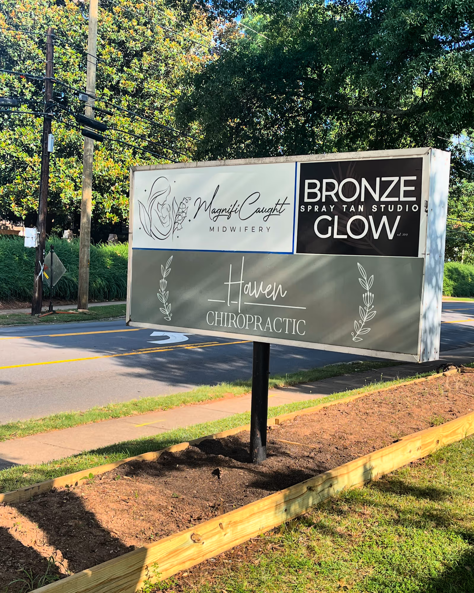 Outdoor signboard displaying MagnifiCaught Midwifery, Bronze Glow, and Haven Chiropractic near road.