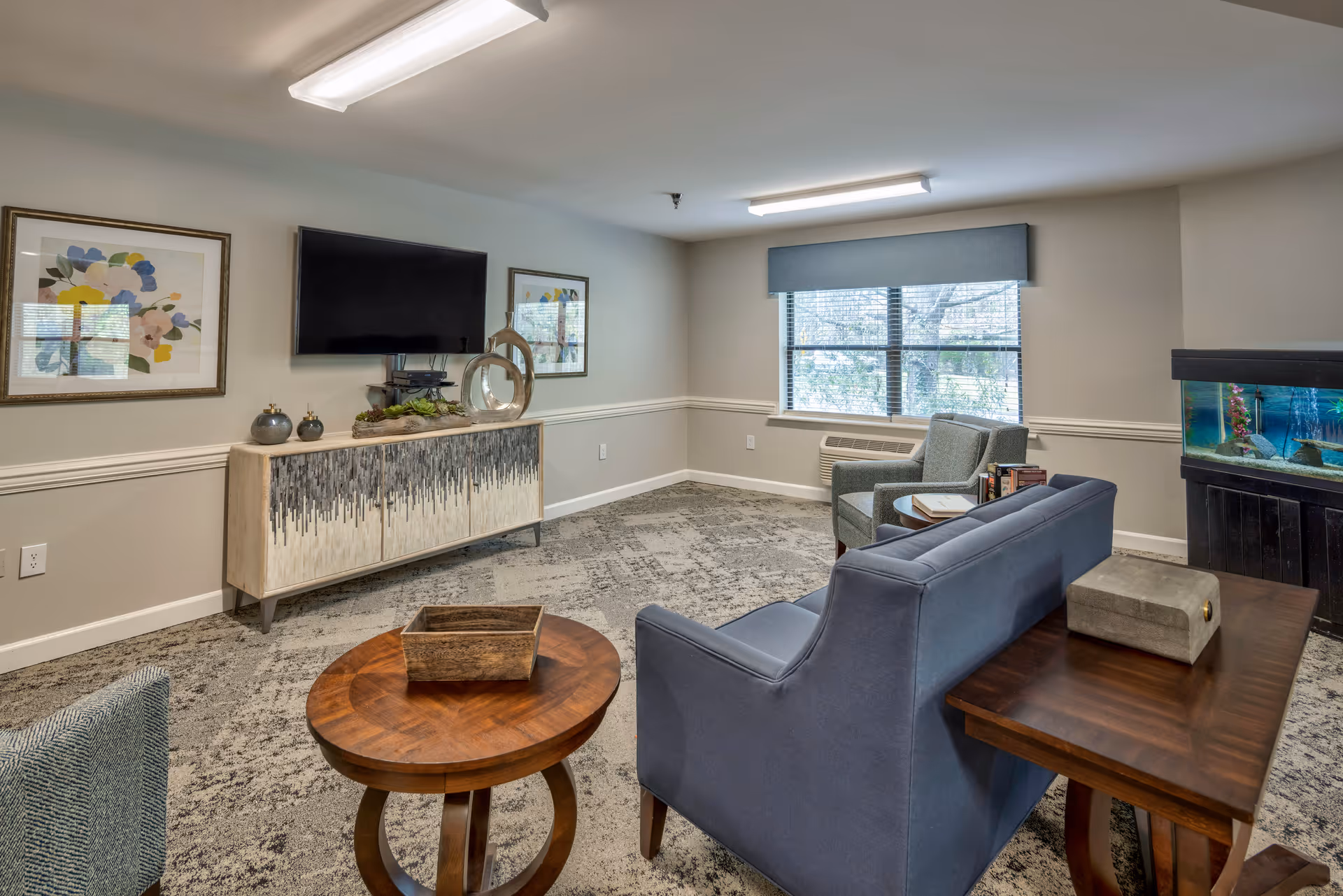 A cozy living room with a blue sofa, two armchairs, a round wooden coffee table, and a wooden side table. A flat-screen TV is mounted on the wall above a decorative cabinet. Two framed floral artworks hang on the wall. A large window with a gray valance lets in natural light. There is also a fish tank on a black stand in the corner of the room.