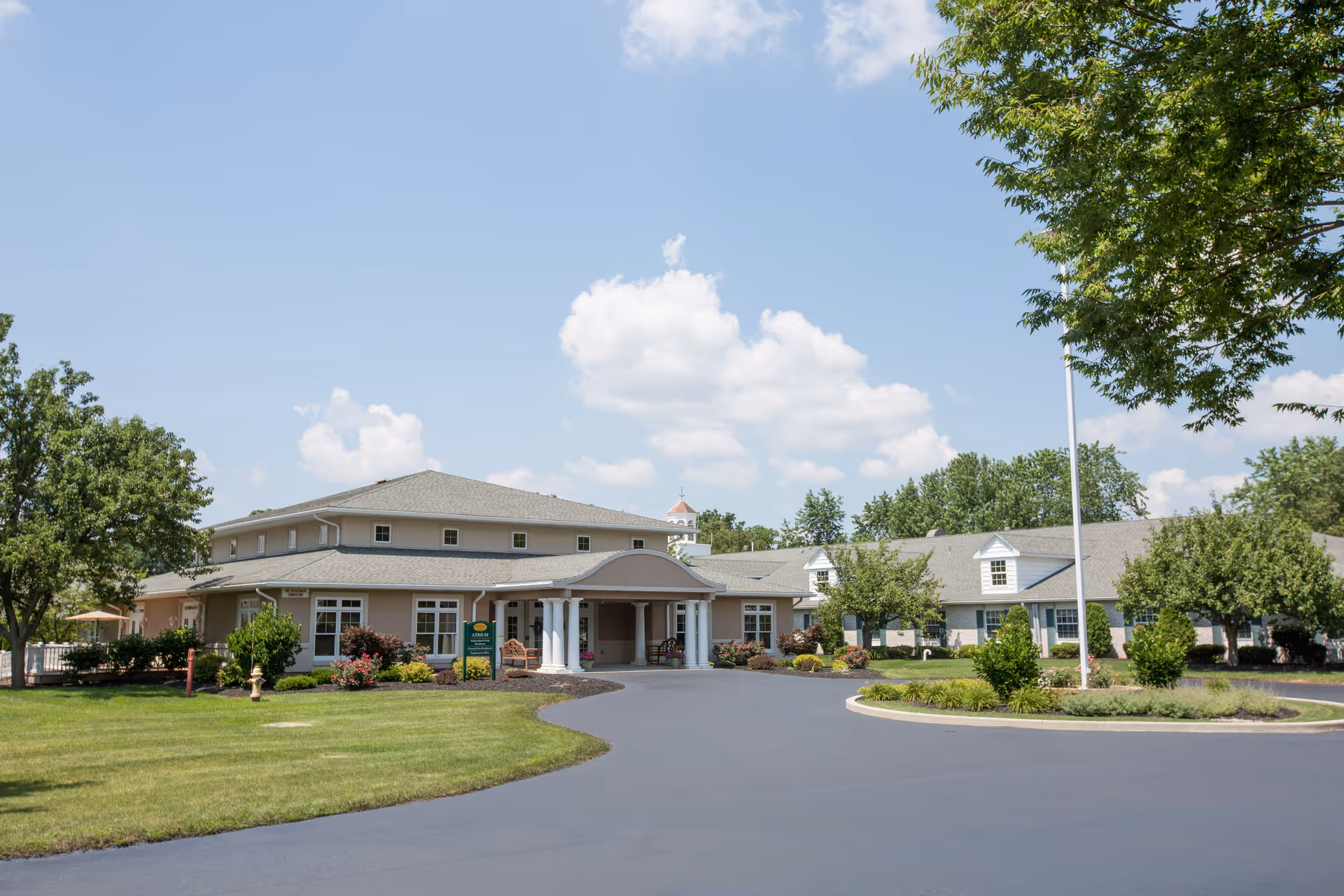 Front exterior of a single-story senior living facility with a circular driveway and landscaped grounds under a blue sky.