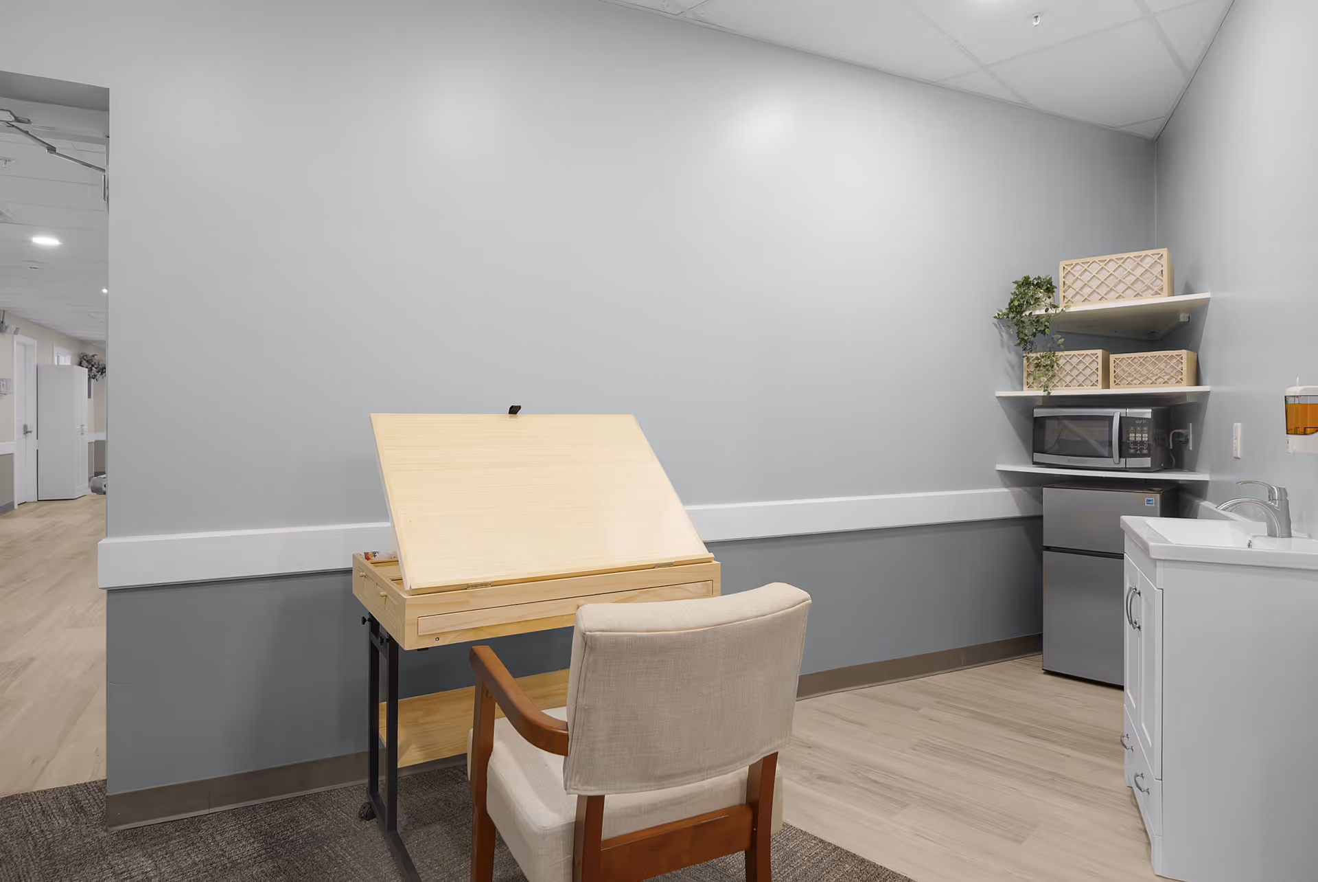 Light blue kitchenette with drafting desk, chair, microwave, mini-fridge, and sink.