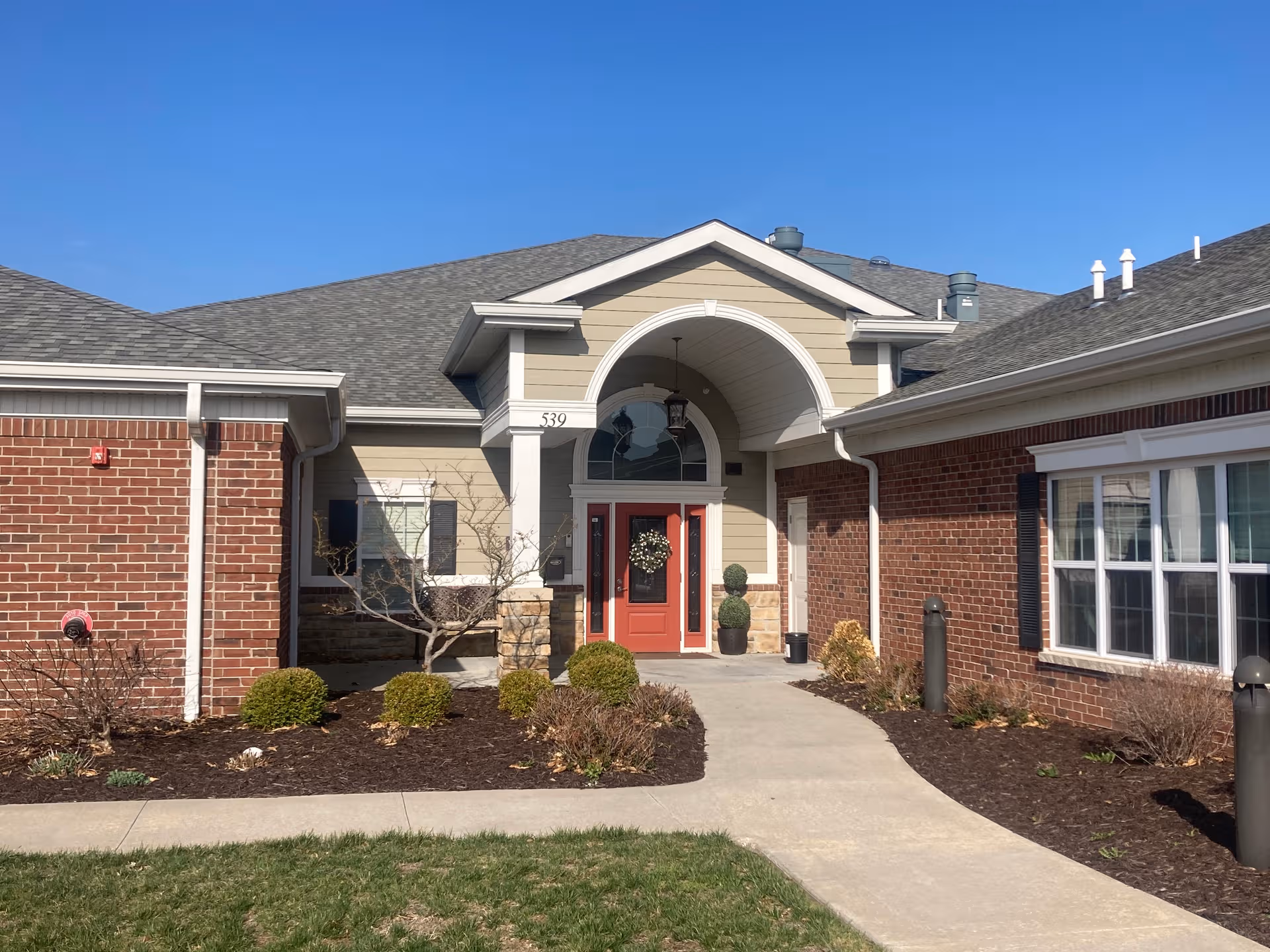 Brick and beige siding senior living facility entrance with red doors and arch.