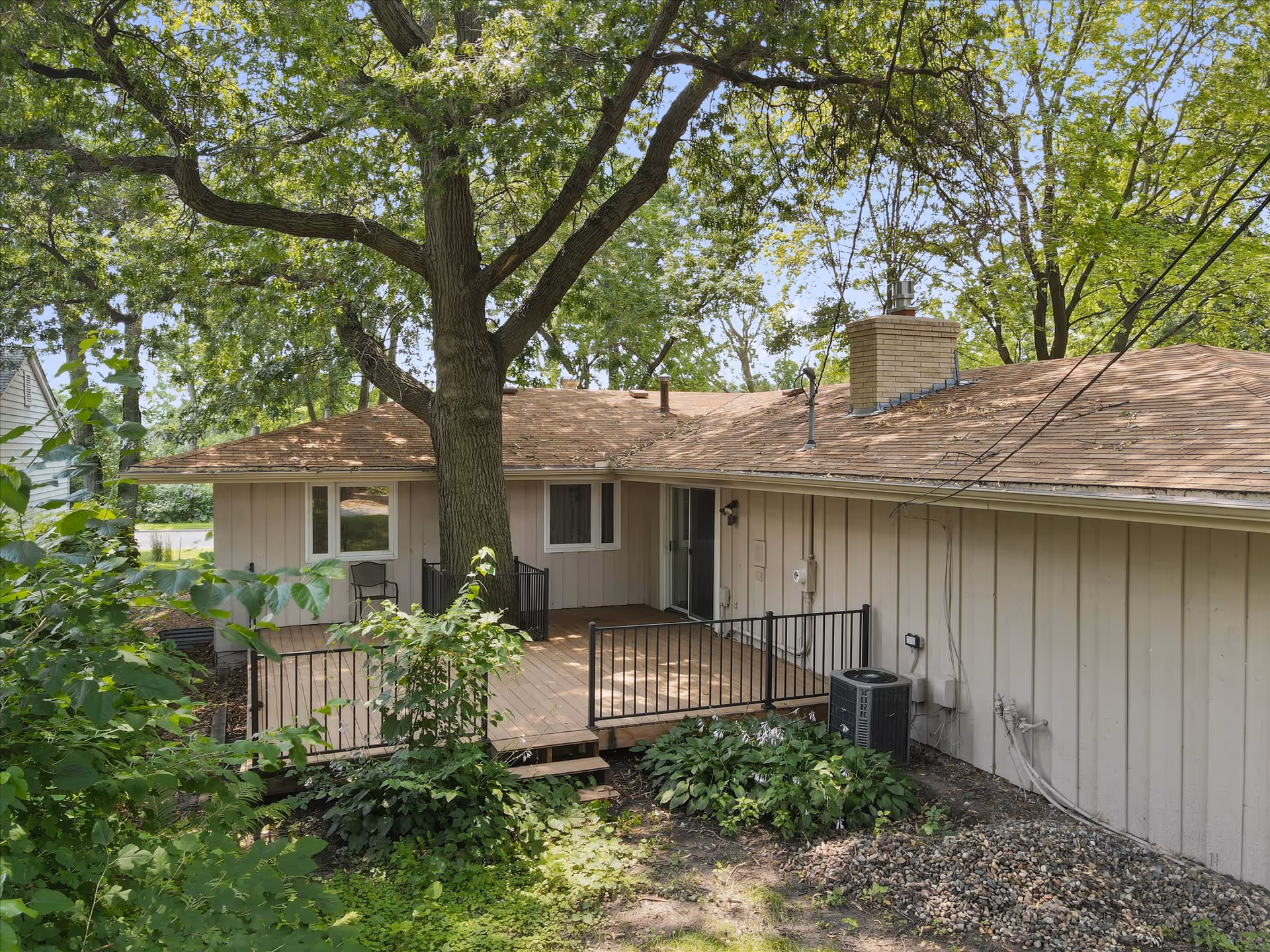 Single-story beige house with wooden deck, railing, and a large tree in a leafy yard.