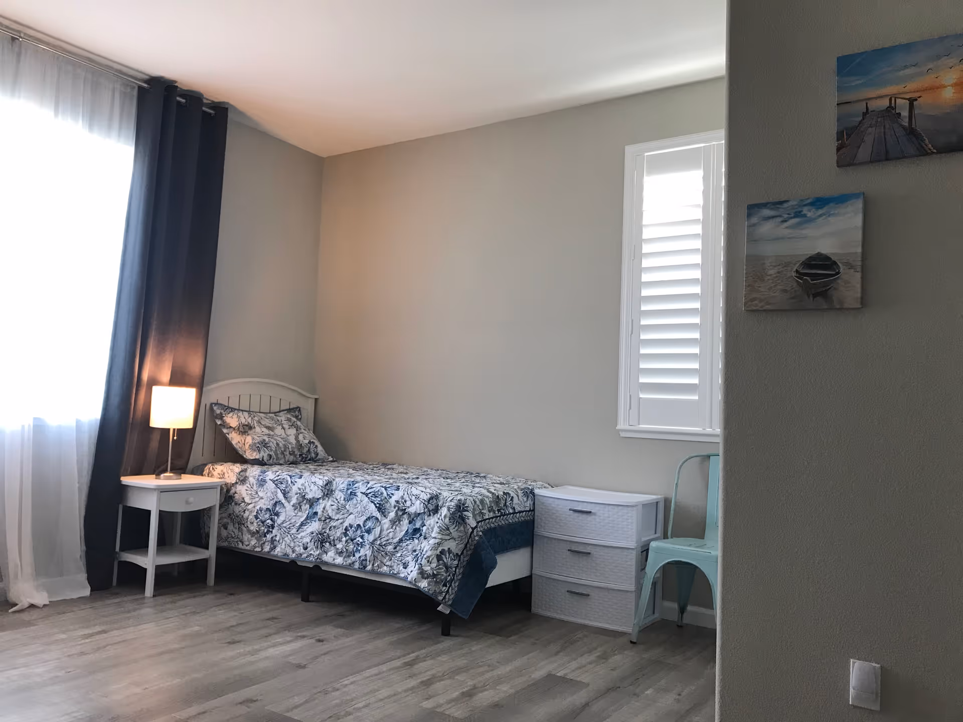 A bedroom in a senior living facility with a single bed, lamp, dresser, and window.